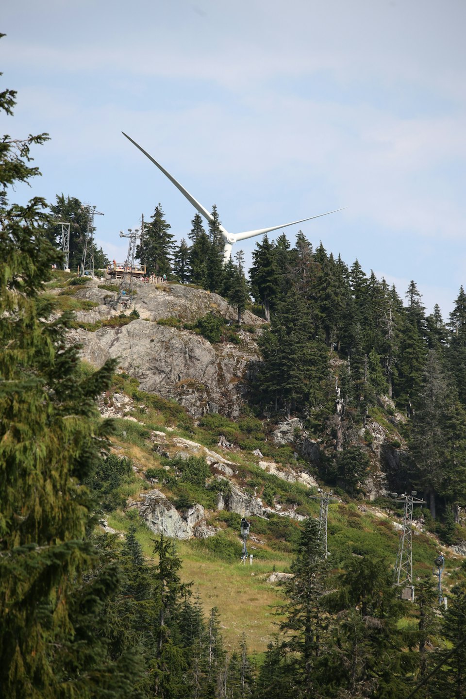 Las aspas de un aerogenerador se alzan en la ladera rocosa y boscosa de una montaña.